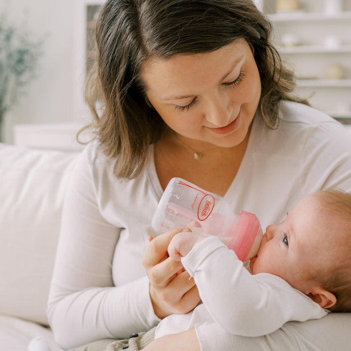 “Mother gently feeding baby with Dr Brown’s anti-colic wide-neck bottle in natural light.